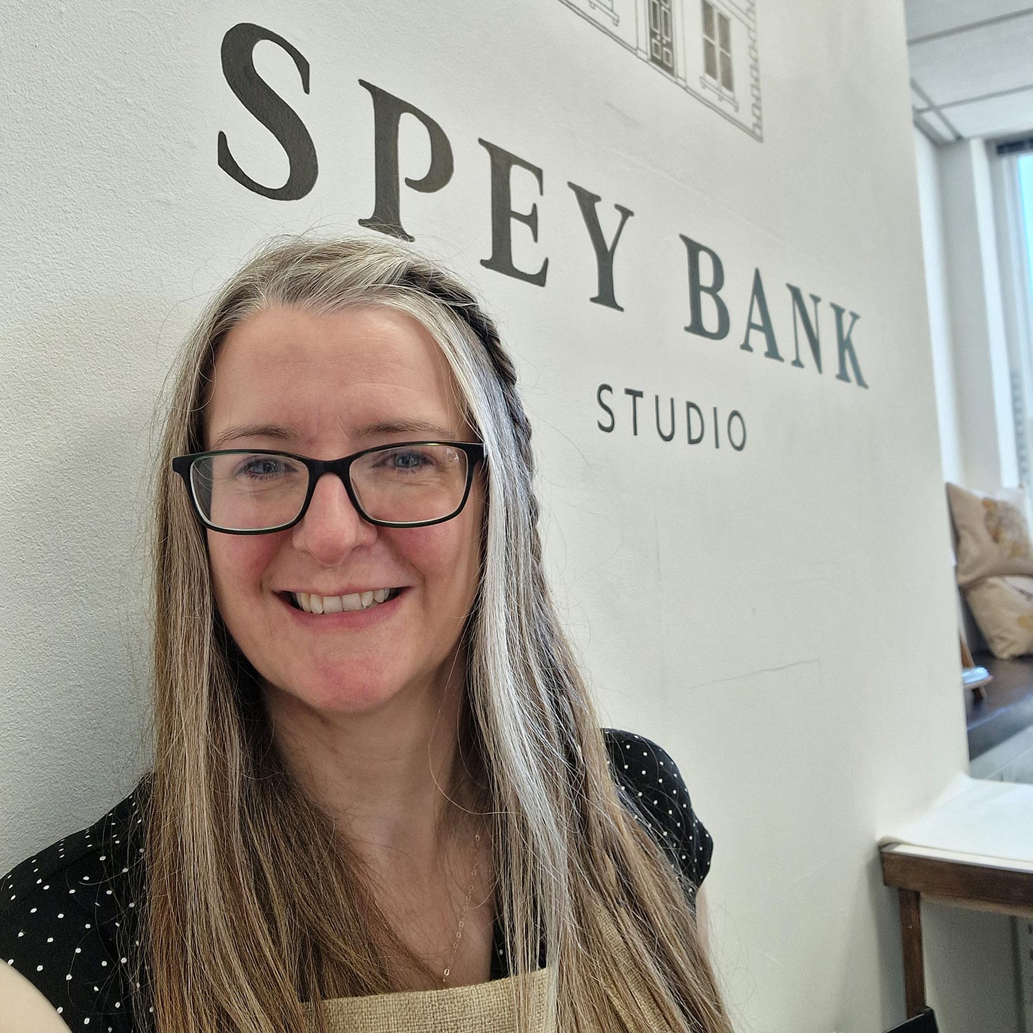 Picture of a woman, Jane Candlish, standing in front of a white wall with the words Spey Bank Studio painted on it.