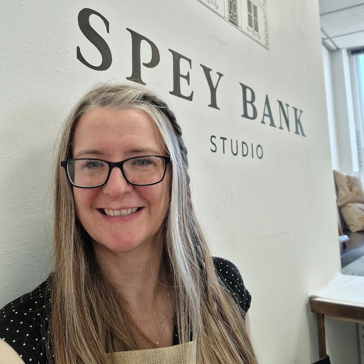 Picture of a woman, Jane Candlish, standing in front of a white wall with the words Spey Bank Studio painted on it.