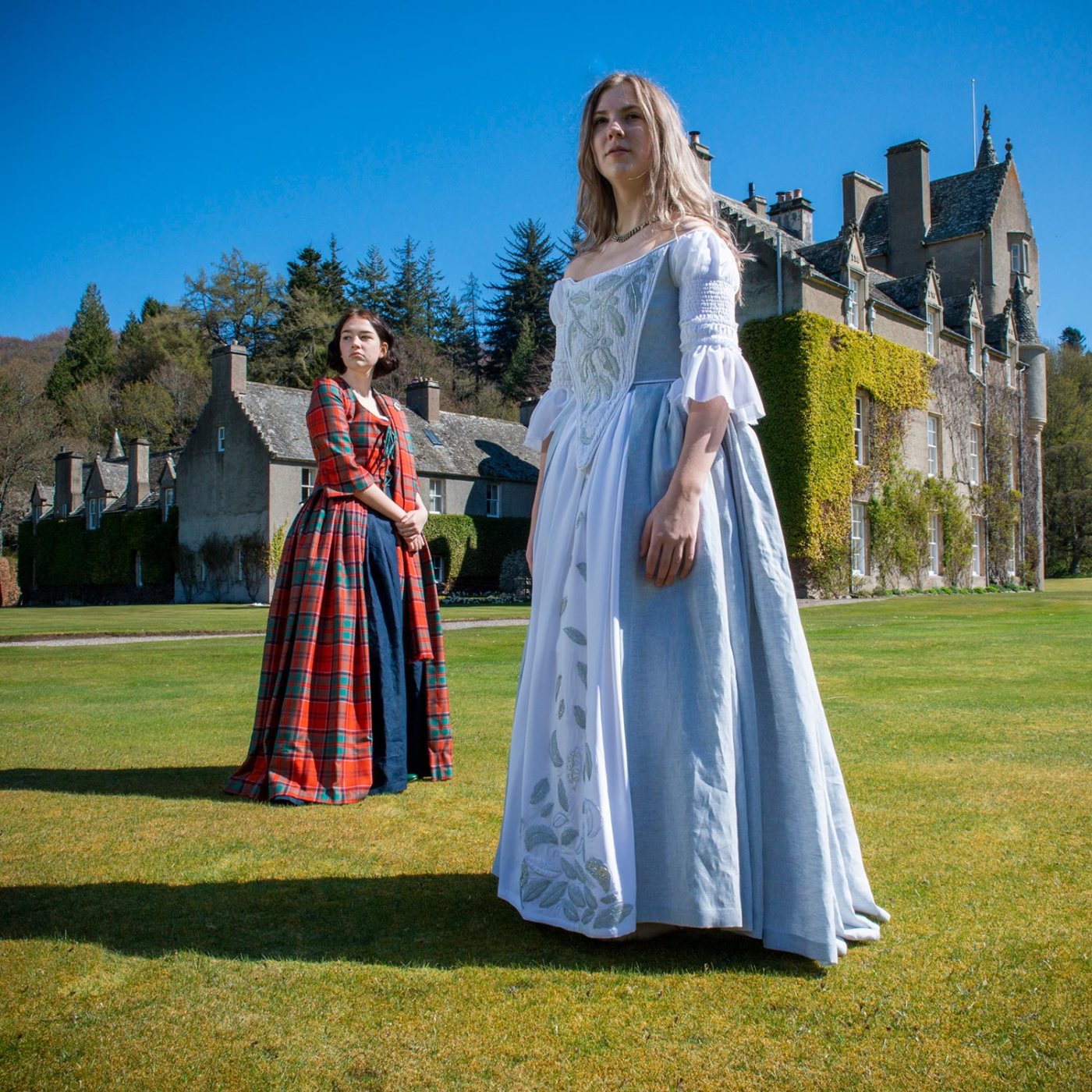 Two women dressed in Jacobite costume, standing on the lawn of a castle on a sunny day