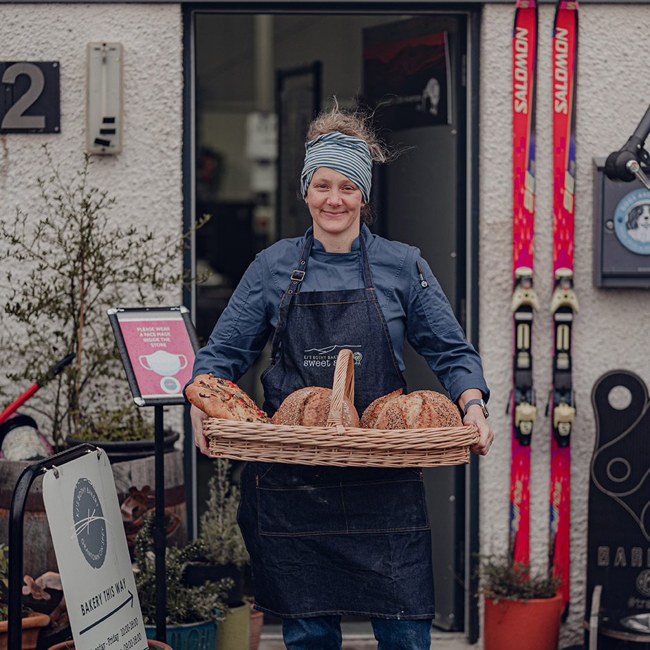A woman, Kirsten Gilmour, holding a basket of bread and standing outside a building. There is a sign for KJ's Bothy Bakery outside and a pair of skis attached to the wall