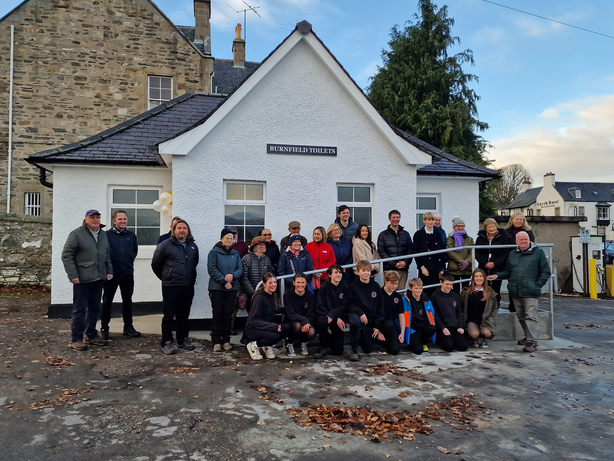 A group of people and teenagers standing outside a small white building. The building has a sign saying Burnfield Toilets.